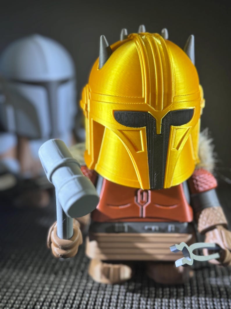 May include: A 3D-printed figure of a character from a science fiction series. The figure has a gold helmet, red and brown armour, and holds a hammer and a tool. Another figure is in the background.