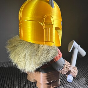May include: A 3D-printed figurine of a character wearing a gold helmet with grey horns, a fur collar, and brown boots. The figure is holding a white and grey hammer. The design is reminiscent of a space warrior.