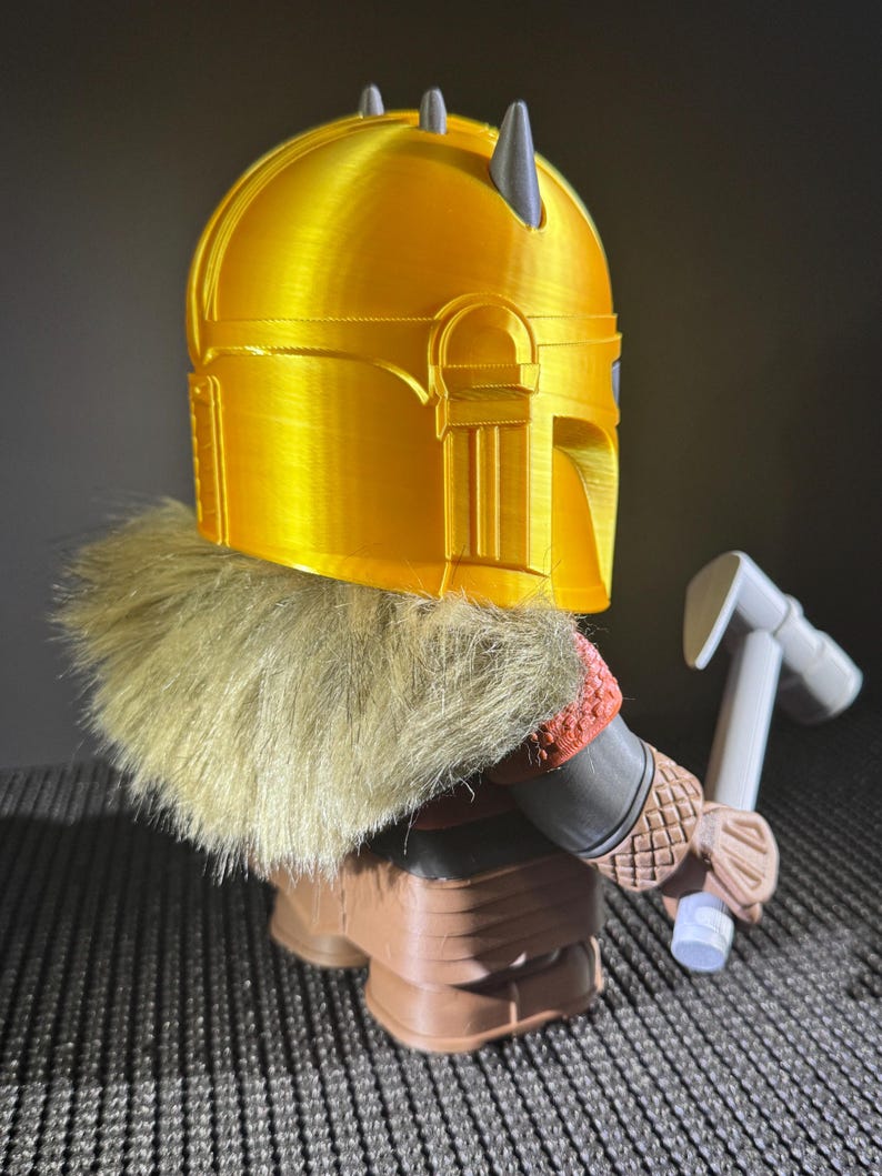 May include: A 3D-printed figurine of a character wearing a gold helmet with grey horns, a fur collar, and brown boots. The figure is holding a white and grey hammer. The design is reminiscent of a space warrior.
