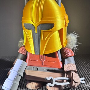 May include: A 3D-printed figure of a character wearing a gold helmet with grey accents and spikes. The figure has a red chest piece, brown arms, and holds a white cylindrical object and a silver tool. The figure is on a grey surface.