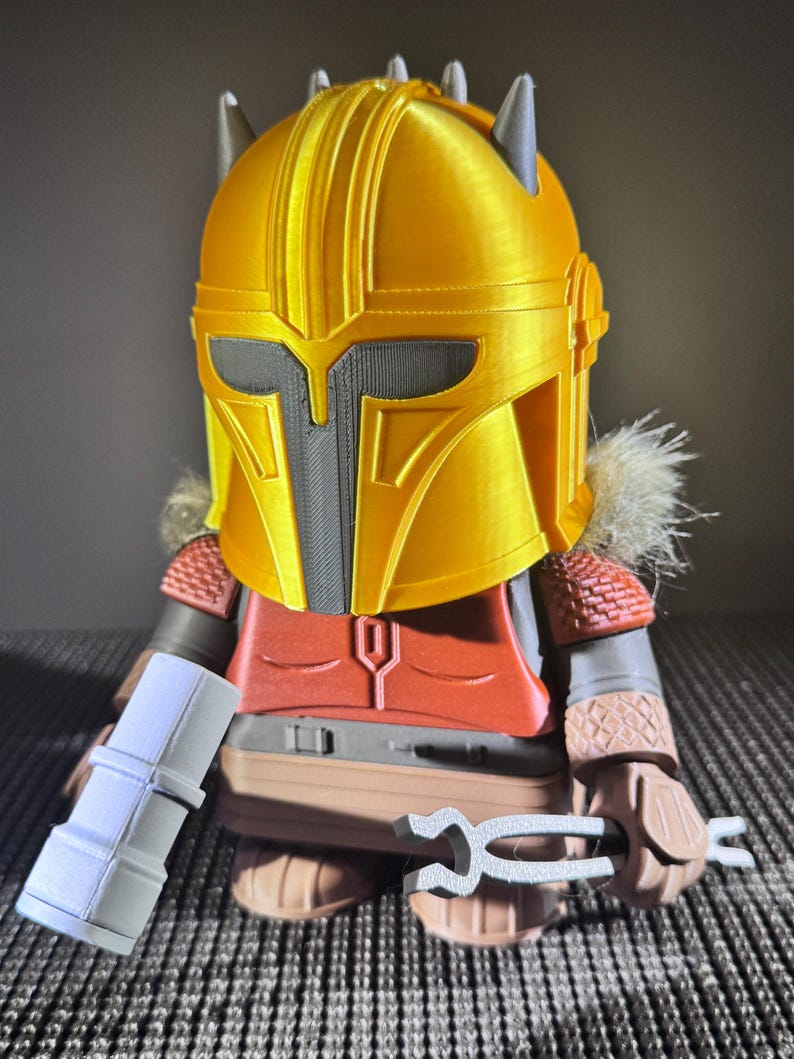 May include: A 3D-printed figure of a character wearing a gold helmet with grey accents and spikes. The figure has a red chest piece, brown arms, and holds a white cylindrical object and a silver tool. The figure is on a grey surface.