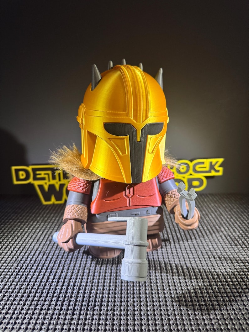 May include: A figurine of a character in a gold helmet, red armour, and a brown fur collar. The figure holds a grey hammer and a small tool. The background is black with yellow text.