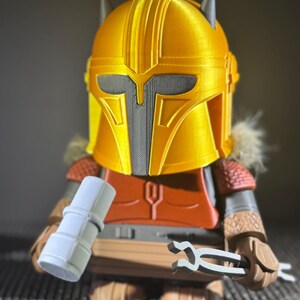 May include: A 3D-printed figure of a character wearing a gold helmet with a grey visor and accents. The figure has an orange chest piece, brown arms, and is holding a grey cylinder and a pair of grey tongs. The figure is a collectible item.