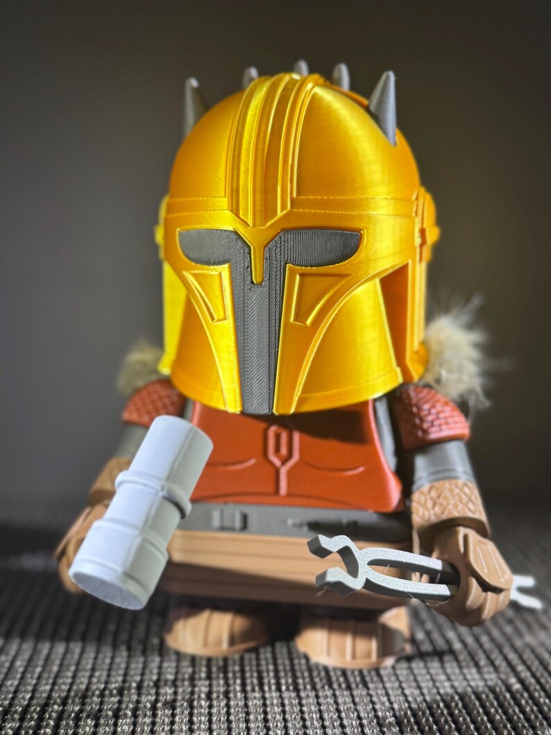 May include: A 3D-printed figure of a character wearing a gold helmet with a grey visor and accents. The figure has an orange chest piece, brown arms, and is holding a grey cylinder and a pair of grey tongs. The figure is a collectible item.