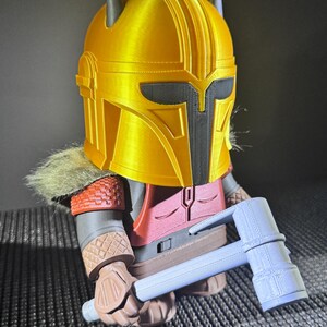 May include: A 3D-printed figurine of a character in golden armour, with a black visor and grey accents. The figure is holding a light blue hammer. The figure has a red and brown outfit with a fur collar.