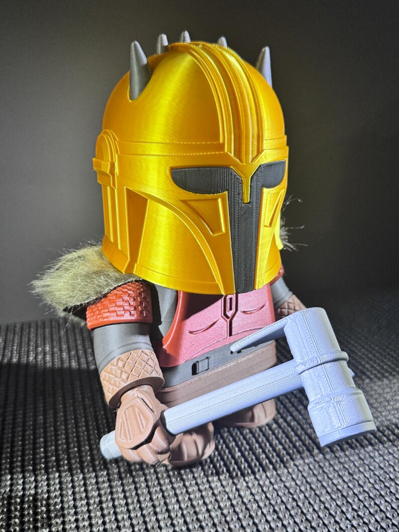May include: A 3D-printed figurine of a character in golden armour, with a black visor and grey accents. The figure is holding a light blue hammer. The figure has a red and brown outfit with a fur collar.