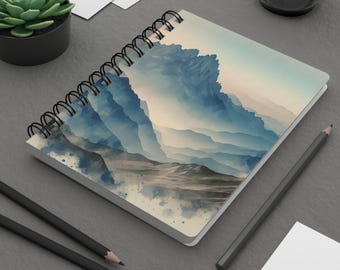 Könnte beinhalten: Ein spiralgebundenes Notizbuch mit einem Aquarellbild einer Bergkette in Blautönen und Grau. Der Hintergrund ist ein weiches, nebliges Blau und die Berge sind mit einem hellbraunen Pfad geschichtet, der sich durch sie hindurchschlängelt.