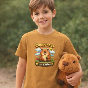K&ouml;nnte beinhalten: Senfgelbes T-Shirt mit dem Text "Cappyness is a Choice" und einer Cartoon-Illustration eines l&auml;chelnden B&auml;ren. Das Kind h&auml;lt ein braunes Stofftier.