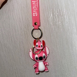 K&ouml;nnte beinhalten: Rosa Stitch-Schl&uuml;sselanh&auml;nger mit passendem Riemen. Der Riemen tr&auml;gt die Worte "Stitch" und "Lovely baby" in Wei&szlig;. Der Schl&uuml;sselanh&auml;nger hat einen goldenen Ring und eine kleine Stitch-Figur auf einer gr&ouml;&szlig;eren Stitch-Figur.
