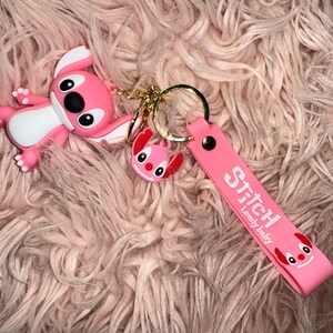 K&ouml;nnte beinhalten: Ein pinkfarbenes Stitch-Schl&uuml;sselanh&auml;nger-Set mit einer gro&szlig;en Stitch-Figur, einem kleineren Stitch-Anh&auml;nger und einem pinkfarbenen Riemen mit dem Text "Stitch Lovely baby". Der Schl&uuml;sselanh&auml;nger hat einen goldfarbenen Ring.