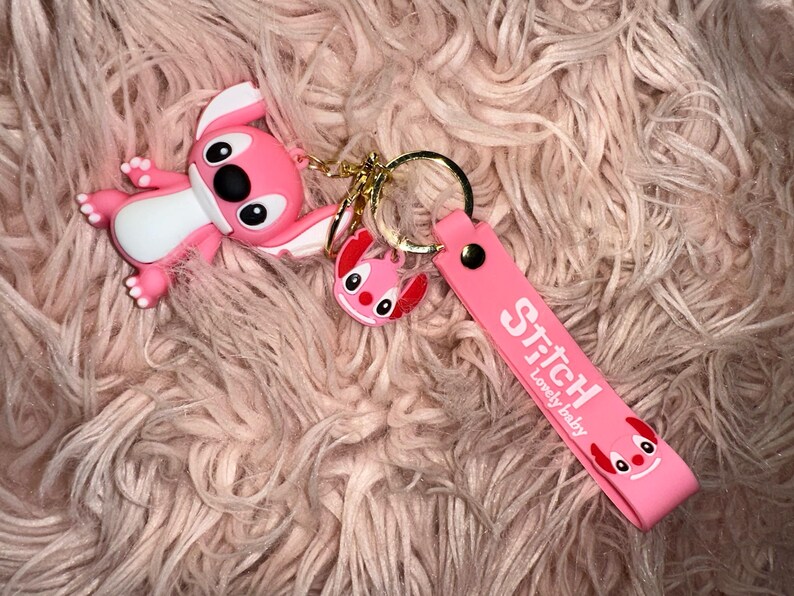 K&ouml;nnte beinhalten: Ein pinkfarbenes Stitch-Schl&uuml;sselanh&auml;nger-Set mit einer gro&szlig;en Stitch-Figur, einem kleineren Stitch-Anh&auml;nger und einem pinkfarbenen Riemen mit dem Text "Stitch Lovely baby". Der Schl&uuml;sselanh&auml;nger hat einen goldfarbenen Ring.