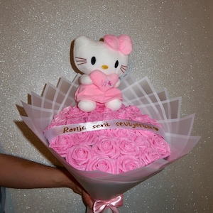Pode incluir: Um buquê de rosas cor-de-rosa embrulhado em papel branco e rosa, coberto com um bicho de pelúcia Hello Kitty segurando um coração. Uma fita com texto dourado envolve as rosas.
