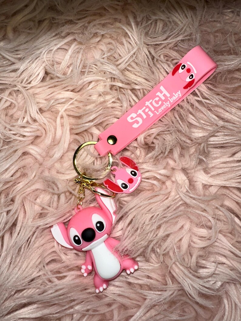 K&ouml;nnte beinhalten: Ein pinkfarbener Stitch-Schl&uuml;sselanh&auml;nger mit einem goldenen Schl&uuml;sselring. Der Schl&uuml;sselanh&auml;nger zeigt eine 3D-Stitch-Figur und einen kleineren Stitch-Kopf-Anh&auml;nger. Auf dem pinkfarbenen Band steht "Stitch Lovely baby" in Wei&szlig;. Der Hintergrund ist eine flauschige, pinkfarbene Oberfl&auml;che.