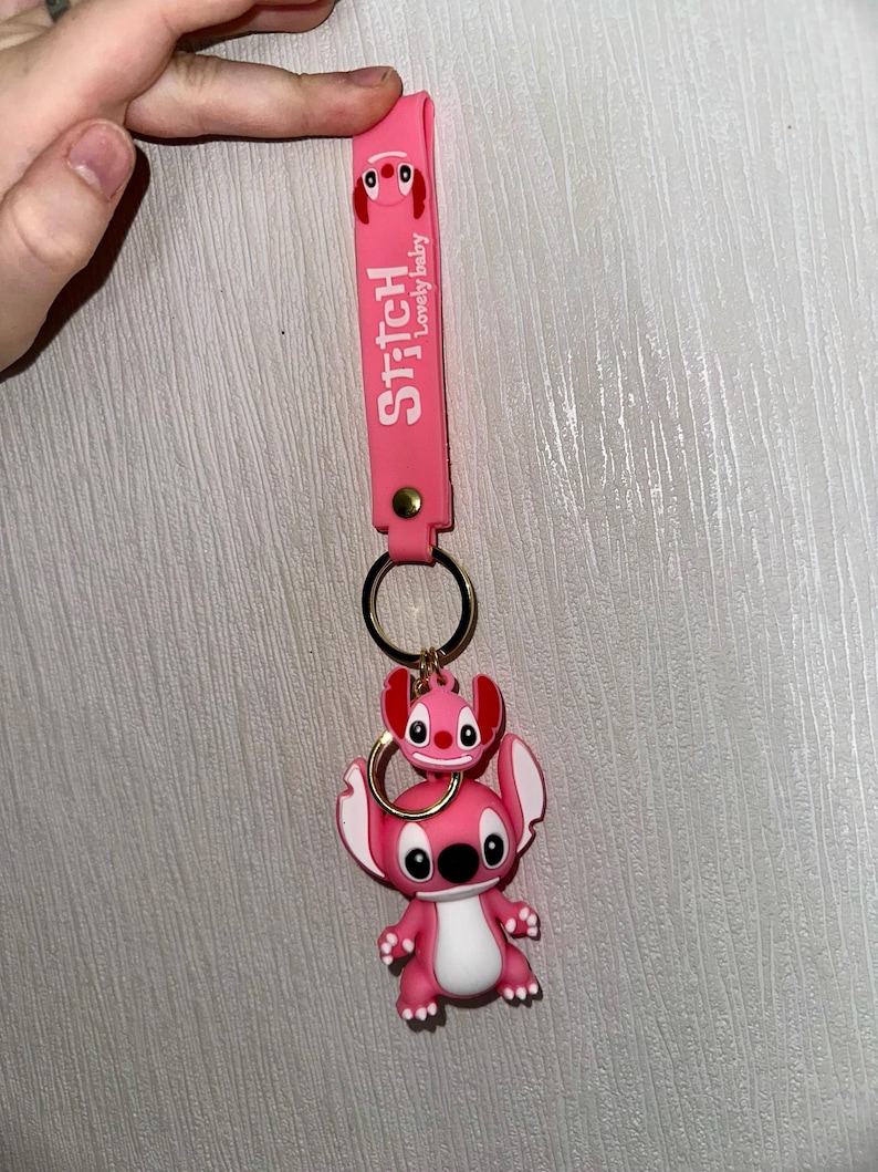 K&ouml;nnte beinhalten: Rosa Stitch-Schl&uuml;sselanh&auml;nger mit passendem Riemen. Der Riemen tr&auml;gt die Worte "Stitch" und "Lovely baby" in Wei&szlig;. Der Schl&uuml;sselanh&auml;nger hat einen goldenen Ring und eine kleine Stitch-Figur auf einer gr&ouml;&szlig;eren Stitch-Figur.