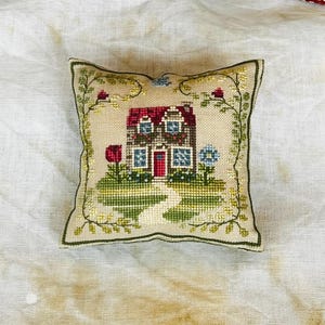 Può includere: Un piccolo puntaspilli quadrato con un disegno a punto croce di una casa a due piani con tetto rosso, porta rossa e finestre blu. La casa è circondata da erba verde, un sentiero bianco e un bordo floreale con fiori rossi e blu.