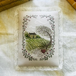 Puede incluir: Un bordado en punto de cruz enmarcado que presenta una escena pastoral con una casa en una colina, un árbol y un corazón. La obra de arte está rodeada por un borde floral y un ribete de encaje. La paleta de colores incluye verde, marrón y rojo.