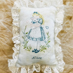 Può includere: Un cuscino rettangolare bianco con un disegno a punto croce di Alice nel Paese delle Meraviglie che tiene un coniglio. Il disegno include dettagli blu e verdi, con il nome "Alice" ricamato sotto. Il cuscino è rifinito con pizzo bianco.