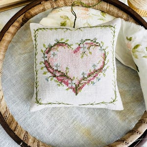 Può includere: Un piccolo cuscino quadrato bianco con un motivo a cuore rosa ricamato a punto croce, adornato con tralci verdi e piccoli fiori blu. Il cuscino è esposto all'interno di un telaio da ricamo in legno.