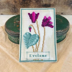 May include: A cross-stitch embroidery of a cyclamen flower with green leaves and a white background. The text "Cyclame" is stitched in a rectangle below the flower.