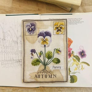 Può includere: Un ricamo a punto croce con un fiore di viola del pensiero con petali viola, gialli e bianchi, foglie verdi e le parole "Pansy" e "Autumn". Il ricamo è su un tessuto beige, con un bordo marrone scuro.