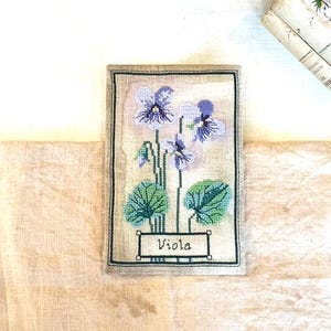 Könnte beinhalten: Eine Kreuzstich-Stickerei einer Veilchenblume mit grünen Blättern und dem Wort "Viola" in einem Rahmen. Die Stickerei ist auf einem beigefarbenen Leinenhintergrund.