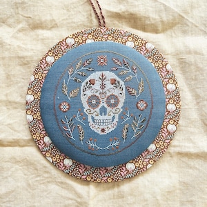 Può includere: Un ornamento rotondo in tessuto blu con un disegno di teschio di zucchero a punto croce. Il teschio è bianco con accenti arancioni e marroni. L'ornamento ha un bordo floreale e un cordino marrone per appenderlo.
