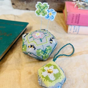 Può includere: Due spilliera fatti a mano con punto ad ago e un tema floreale. Lo spilliera più grande ha la forma di un esagono e presenta un fiore bianco con foglie verdi, un'ape e un trifoglio a quattro foglie. Lo spilliera più piccolo ha la forma di una lacrima e presenta un fiore bianco con foglie verdi e un'ape.