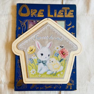 Può includere: Un ornamento da parete a forma di casa ricamato a punto croce, con un coniglietto e un pulcino in un campo di fiori, con la scritta "Sweet home". Su un libro blu con lettere dorate.