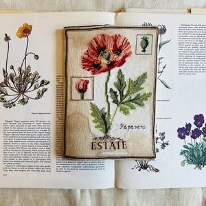 Può includere: Ricamo a punto croce di un fiore di papavero rosso con foglie verdi e la parola "Estate" sotto. Il disegno è su un tessuto beige, con la parola "Papavero" sopra il fiore. Il ricamo è posto su un libro aperto.
