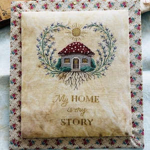 Può includere: Ricamo a punto croce raffigurante una casa con tetto a fungo rosso e bianco, circondata da una ghirlanda verde e blu e un sole. Il testo "My HOME is my STORY" è ricamato sotto. L'opera è incorniciata con tessuto floreale.
