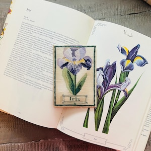 Può includere: Un segnalibro ricamato a punto croce con un fiore di Iris con bordo verde, appoggiato su un libro aperto con illustrazioni botaniche. Il segnalibro presenta un disegno di Iris viola e giallo con la parola "Iris" sotto.