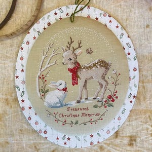 Può includere: Ornamento natalizio ovale con un disegno a punto croce raffigurante un cervo con una sciarpa rossa e un coniglio bianco. L'ornamento ha uno sfondo color crema e il testo "Treasured Christmas Memories". Il bordo è bianco con stelle e palline rosse.