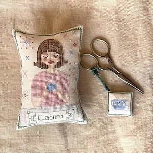 Può includere: Un cuscino bianco in punto ago con un disegno a punto croce di una ragazza che tiene una tazza. Il cuscino ha un bordo verde e il testo "Laura" è ricamato in basso. È anche raffigurato un piccolo quadrato bianco in punto ago con un disegno a punto croce blu, insieme a un paio di forbici argentate.