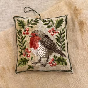 Può includere: Ornamento quadrato a punto croce con un'immagine dettagliata di un pettirosso. L'uccello ha il petto rosso, la testa grigia e le ali marroni, circondato da foglie verdi e bacche rosse. L'ornamento è incorniciato con un bordo verde e ha un anello verde scuro per appenderlo.