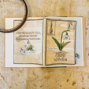Può includere: Libro aperto con illustrazioni a punto croce di bucaneve. Una pagina presenta il testo "Amid the winter's frost, snowdrops whisper that nothing truly ends." L'altra pagina mostra una pianta di Galanthus con la parola "WINTER".