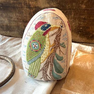 Può includere: Un cuscino ricamato a mano con un uccello colorato appollaiato su un ramo. L'uccello ha un corpo verde, un becco giallo e una testa rossa. Lo sfondo è in tessuto di lino color crema.