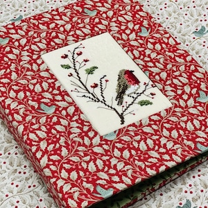 Può includere: Una scatola a fantasia rossa e bianca con un'immagine ricamata a punto croce di un uccello su un ramo. La scatola è ricoperta da un motivo a agrifoglio e bacche. Il punto croce presenta un uccello dal petto rosso, foglie verdi e bacche rosse.