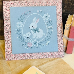 Può includere: Un ricamo a punto croce incorniciato con un coniglio bianco con un fiocco blu, un piccolo uccello e una ghirlanda di fiori su uno sfondo blu chiaro.