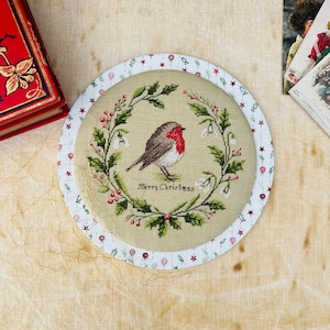 Può includere: Una decorazione natalizia ricamata, di forma circolare, con un motivo a punto croce di un pettirosso, circondato da una ghirlanda di verde e bacche. Le parole "Merry Christmas" sono ricamate sotto. Il bordo esterno è color crema con un motivo di stelle rosse e piccole palle.