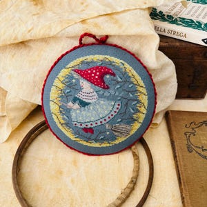 Può includere: Ricamo a punto croce circolare raffigurante una strega con un cappello rosso a pois bianchi, su una scopa. Il disegno è racchiuso in un bordo di foglie gialle e verdi su sfondo blu, incorniciato con filo rosso.