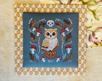 May include: A cross-stitch design featuring an owl perched on a pumpkin, surrounded by mushrooms and autumn leaves, with a skull above the owl. The design is on a teal background, framed by a gold floral pattern. The text "31st" is at the bottom.