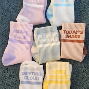 May include: Seven pairs of ribbed crew socks in various pastel colours, each featuring a horizontal stripe and text including RAINBOW SOUL, OCEAN TIDE, HAPPY FACE, PLAYLIST SHUFFLE, TODAY'S SHADE, DRIFTING CLOUD, and MIND MOSAIC printed on the cuffs.