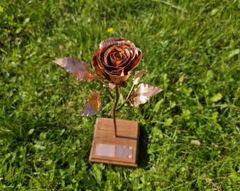 Puede incluir: Una escultura de rosa de metal hecha a mano con pétalos y hojas de color cobre. La rosa está montada sobre una base de madera con una pequeña placa de metal. La escultura está sobre un fondo de hierba verde.