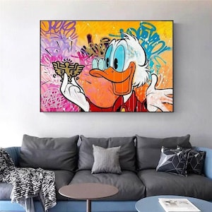 Puede incluir: Una impresión enmarcada de arte pop que presenta un personaje de pato de dibujos animados con cabeza blanca, pico naranja y ojos azules. La obra de arte tiene un fondo de estilo graffiti con colores vibrantes y un objeto dorado en la mano del pato.