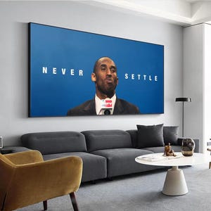May include: A framed wall art print featuring a portrait of a man in a suit against a blue background. The words "NEVER SETTLE" are printed in white on the blue background. The art is displayed above a gray sofa in a modern living room.
