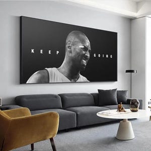 May include: Black and white wall art featuring a portrait of a basketball player with the words "KEEP GOING". The artwork is displayed above a gray sofa in a modern living room setting.