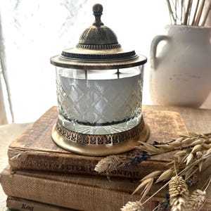 May include: A vintage-style candle holder with a glass jar and a bronze-coloured lid and base. The glass jar has a diamond pattern. The candle holder is on a stack of old books. Dried flowers and a white vase are in the background.