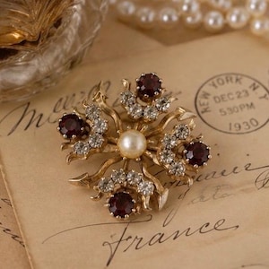 May include: A gold-tone metal brooch featuring a central cream-colored faux pearl surrounded by four deep red circular stones and clusters of small clear rhinestones arranged in a star-like pattern with curved, pointed metal arms extending outward.