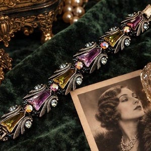 May include: A link bracelet featuring alternating purple and yellow faceted stones set in an antiqued metal frame. The design includes smaller clear and iridescent round crystals accented by textured metal leaf-like details and a secure clasp.