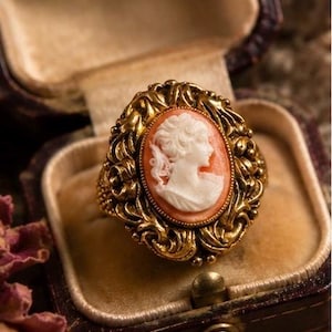 May include: An oval cameo ring featuring a white profile portrait of a person carved against a peach-colored background. The central cameo is set within an ornate, textured gold-tone metal frame with intricate scrollwork and decorative detailing.